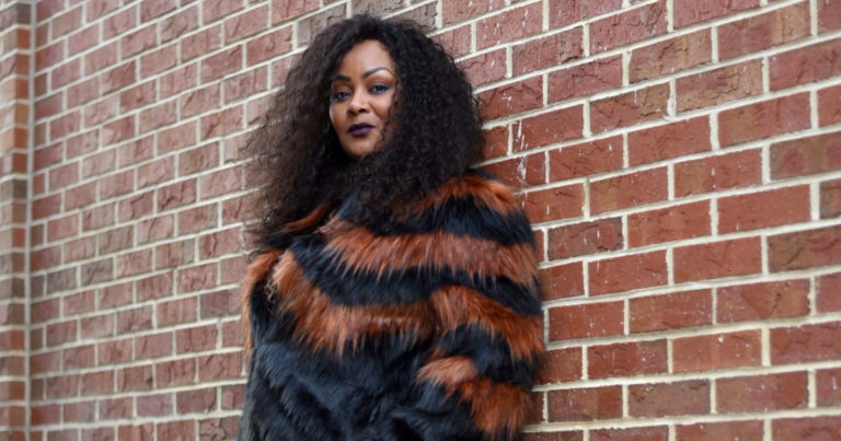 plus size woman in faux fur jacket leaning against brick wall